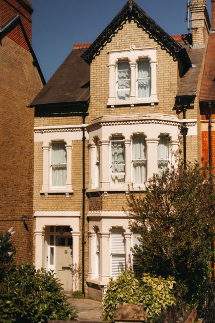 A photograph of Belsize Park station in NW3, showing a large, historic brick building with arched windows and a central clock feature, situated behind a spacious open area with parked cars, planters,